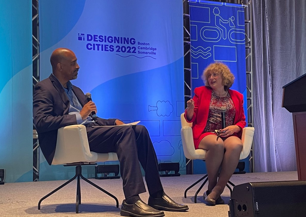 A Black man in a dark navy suit holds a microphone while listening to a curly-haired white woman in a red suit while she gestures with one hand and holds a microphone in the other. They are sitting on a stage in a conference room in front of a light blue banner that reads "NACTO Designing Cities 2022"