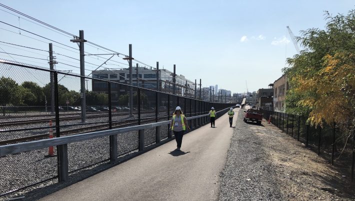 Three people in construction vests walk down a long, straight paved pathway next to railroad tracks. In the distance the path ascends a ramp then curves out of sight, and beyond that is the skyline of downtown Boston.