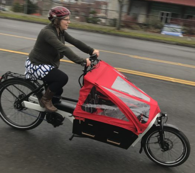 A woman wearing a sweatshirt, patterned skirt, and bike helmet pedals a battery-powered e-bike with a large cargo compartment that's covered in a red fabric tent in front of her handlebars.