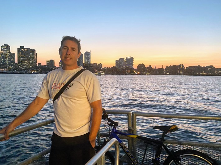 Greg poses next to his bike on the outdoor part of the ferry with the Boston skyline in the background
