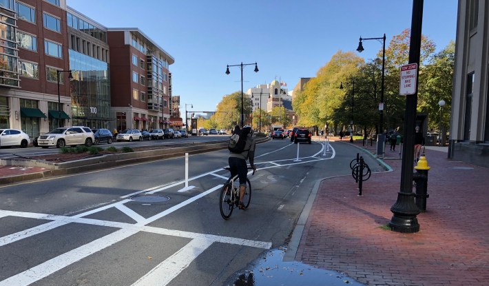 A bicyclist rides in a bike lane between a sidewalk and a row of white flexible-post bollards. A sign on a lampost in the foreground reads "tow zone - no stopping bike lane." In the distance, cars wait at a red light.