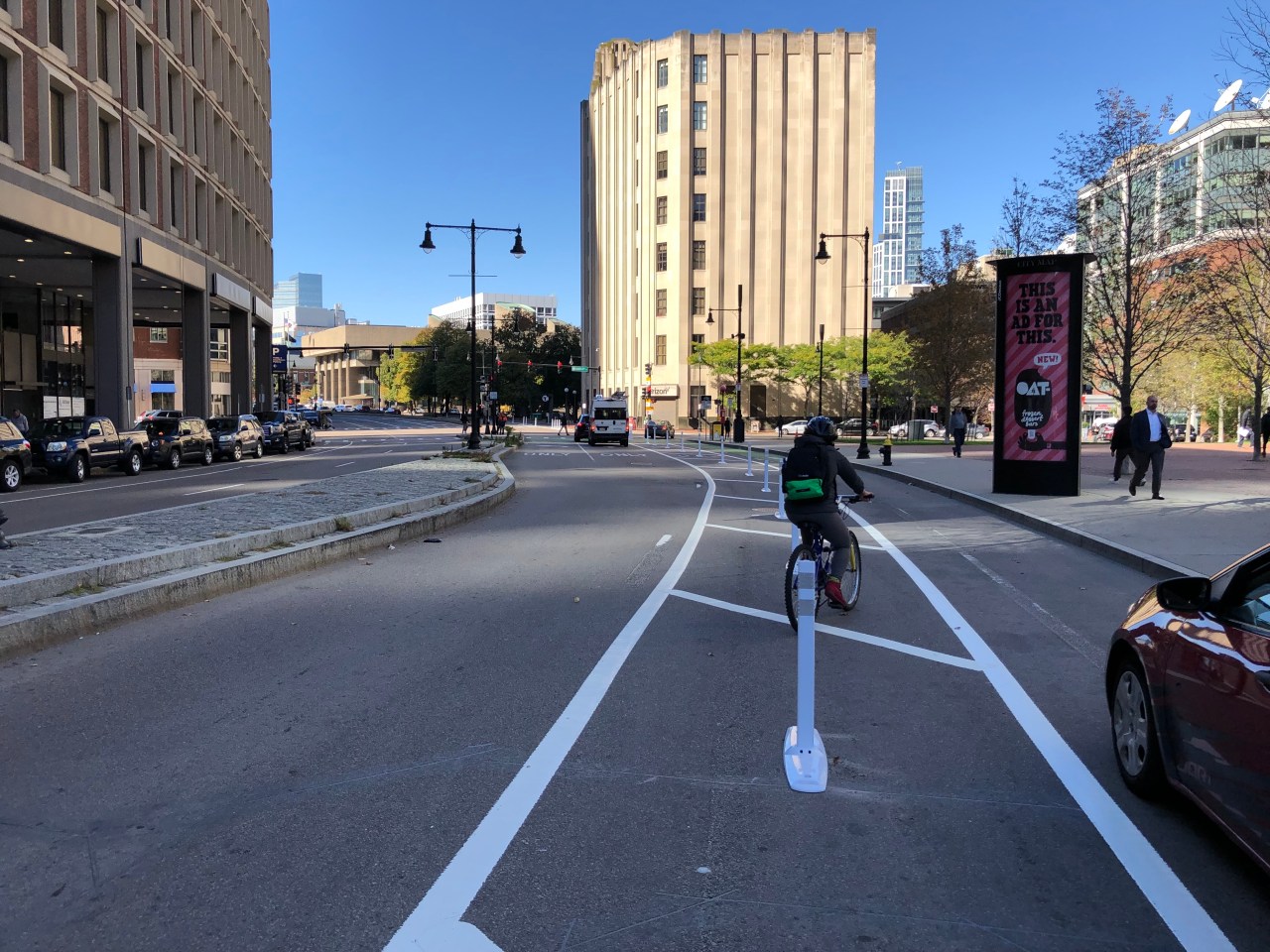 A bicycle user enters a bike lane in front of an illegally-parked car along City Hall Plaza in downtown Boston next to an otherwise-empty street. In the distance, two cars wait an a red traffic light.