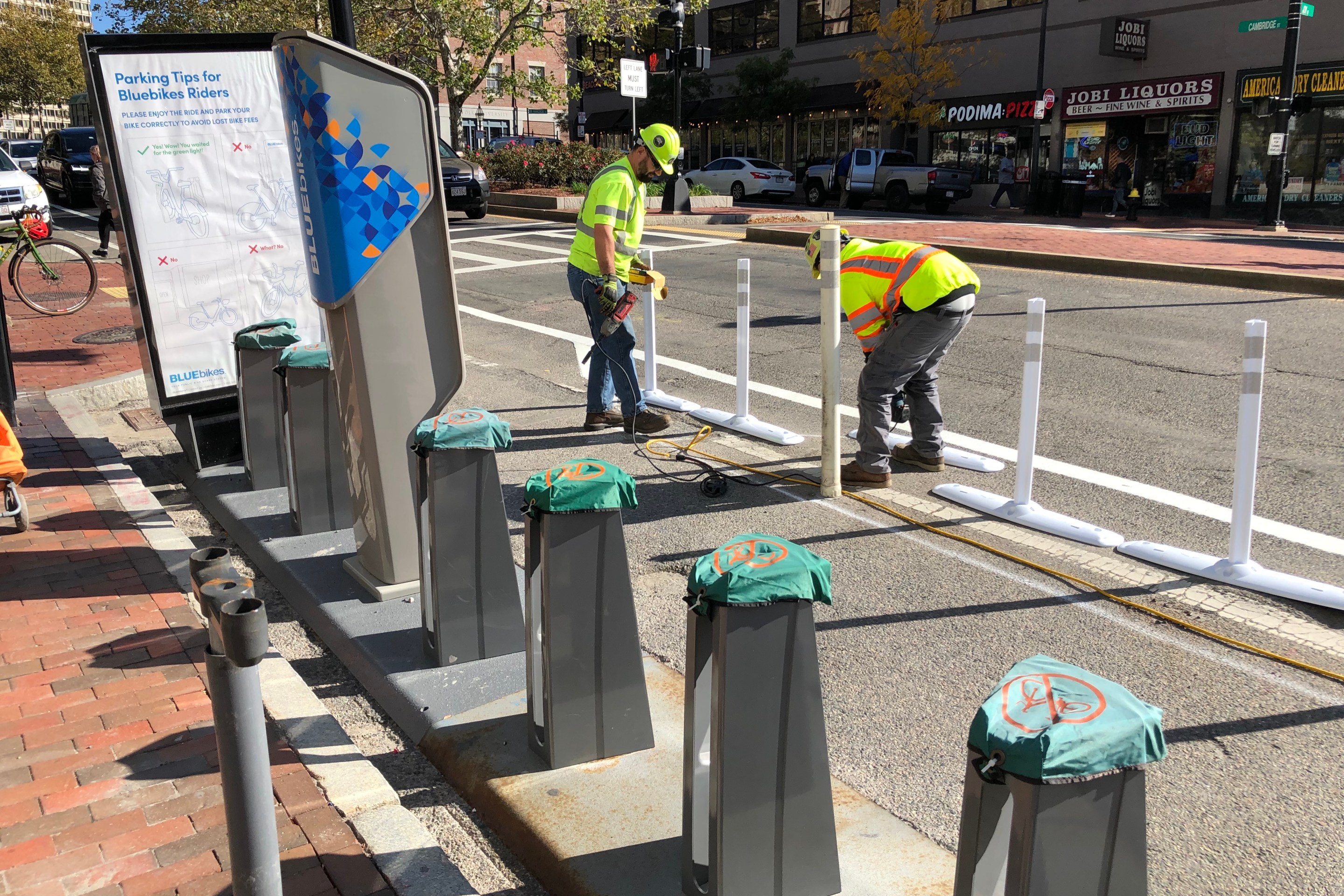 Boston Transportation Department workers in bright yellow vests install flexible-post bollards for the new Cambridge Street bike lane next to an empty Bluebikes dock.