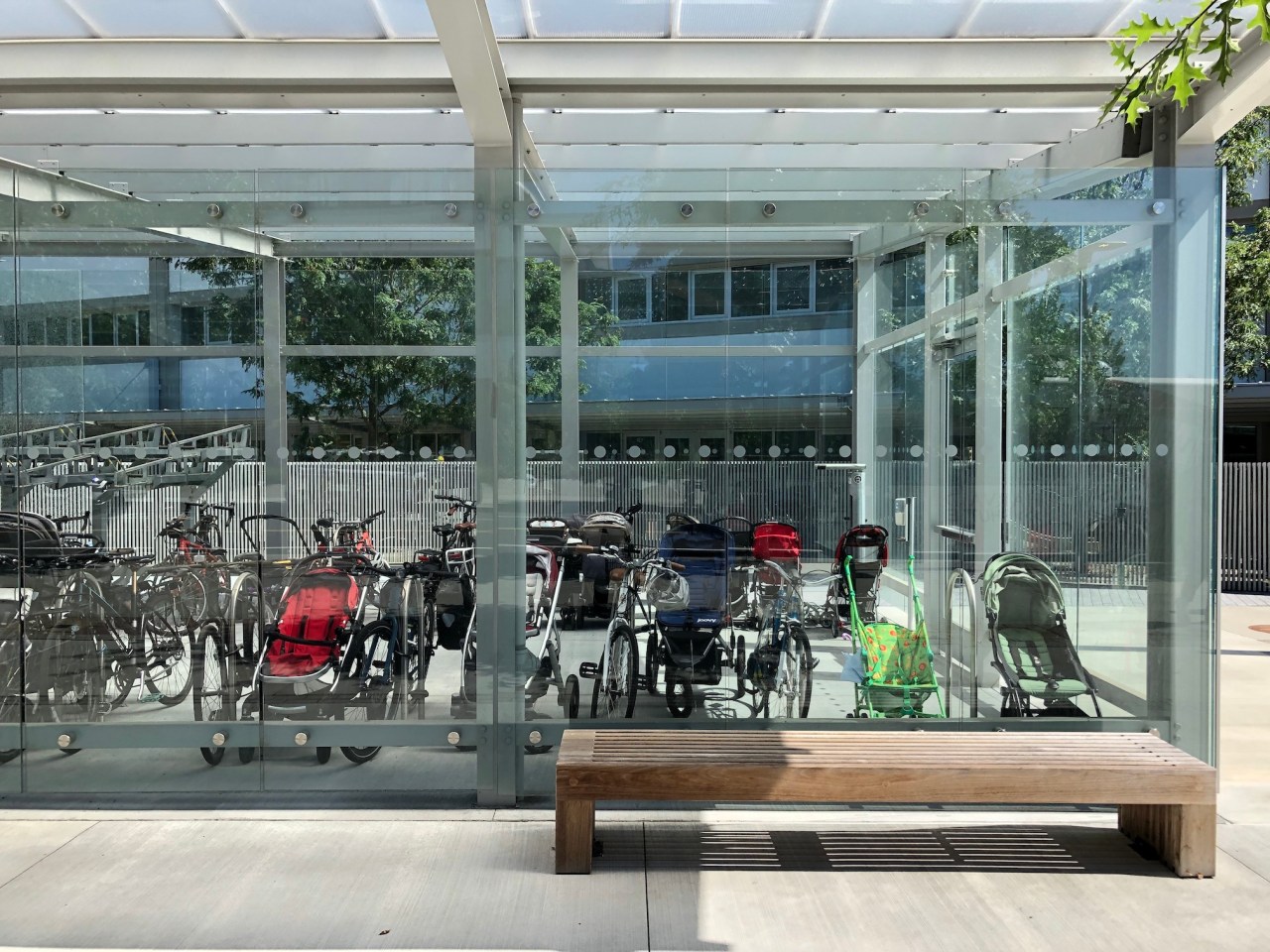 About a dozen baby strollers stored inside a glass-clad bike parking shed next to bike racks.