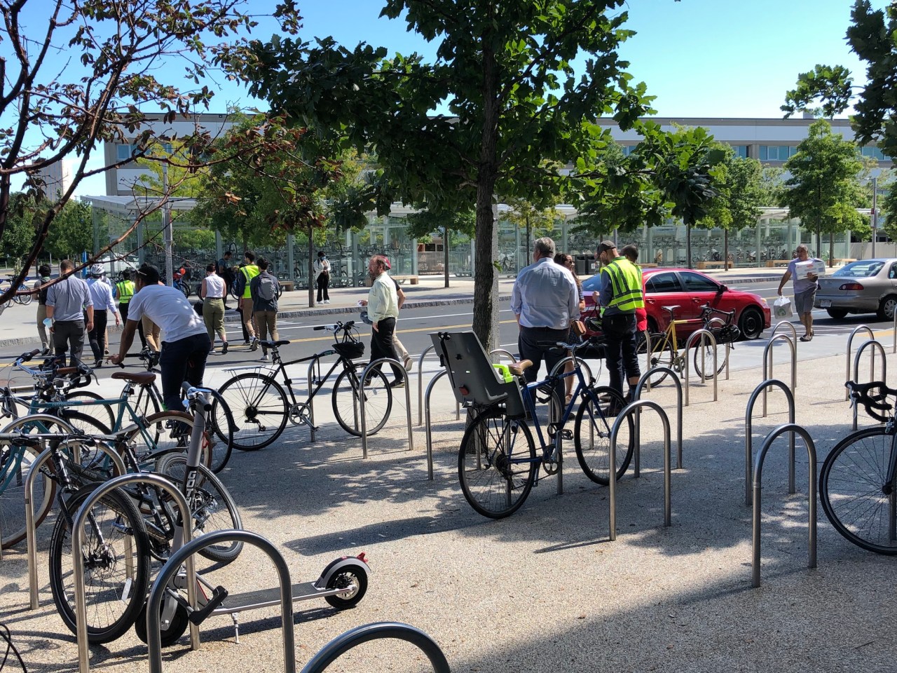 An array of U-shaped ground-mounted bike racks occupies a courtyard next to a small street. In the distance, on the other side of the street, is a long glass-clad bike parking shed full of bikes..