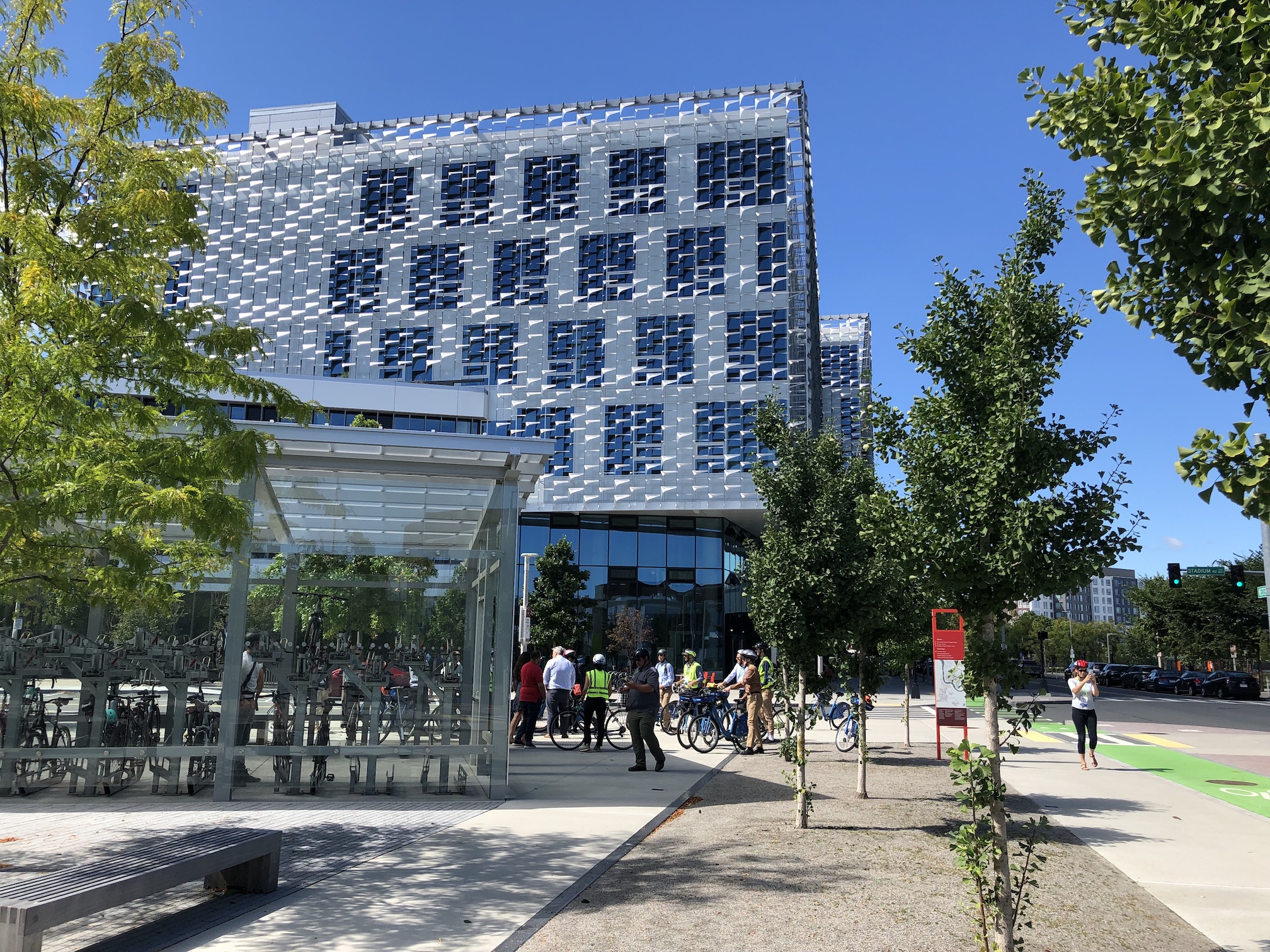 An exterior photo of the new Harvard Engineering School building, next to a protected bike lane. A large group of people on bikes is gathered in the foreground next to a glass-clad bike parking shed while a jogger runs by on the sidewalk.