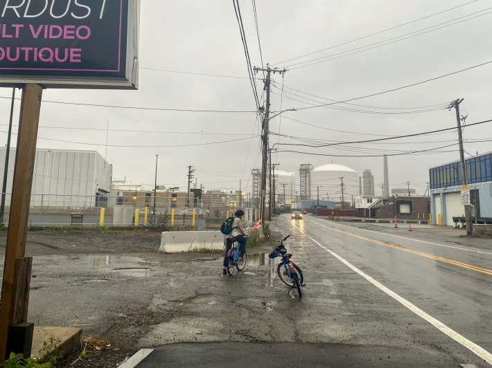 a bicyclist waits to cross the road on a muddy stretch of degrading asphalt on a rainy day
