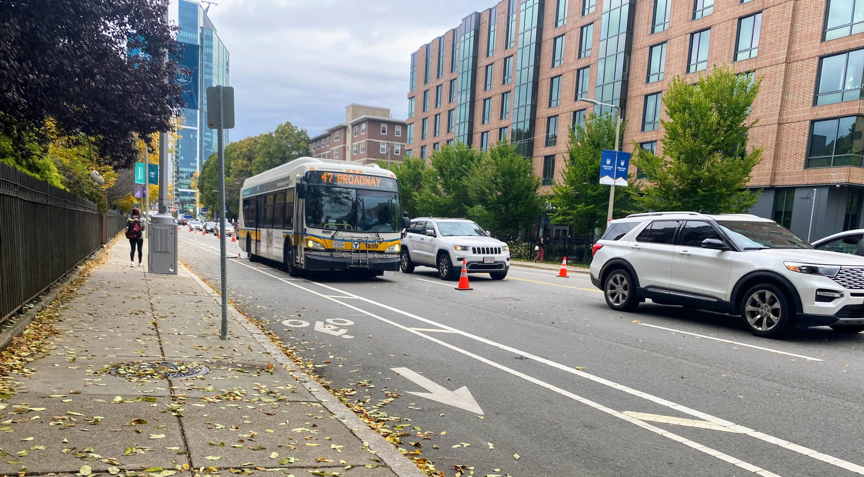 A black-and-yellow MBTA route 47 bus drives in an empty lane delineated by a row of cones from an adjacent car lane where multiple cars and SUVs are stuck in traffic. To the left of the bus, along the curb, is a bicycle lane.