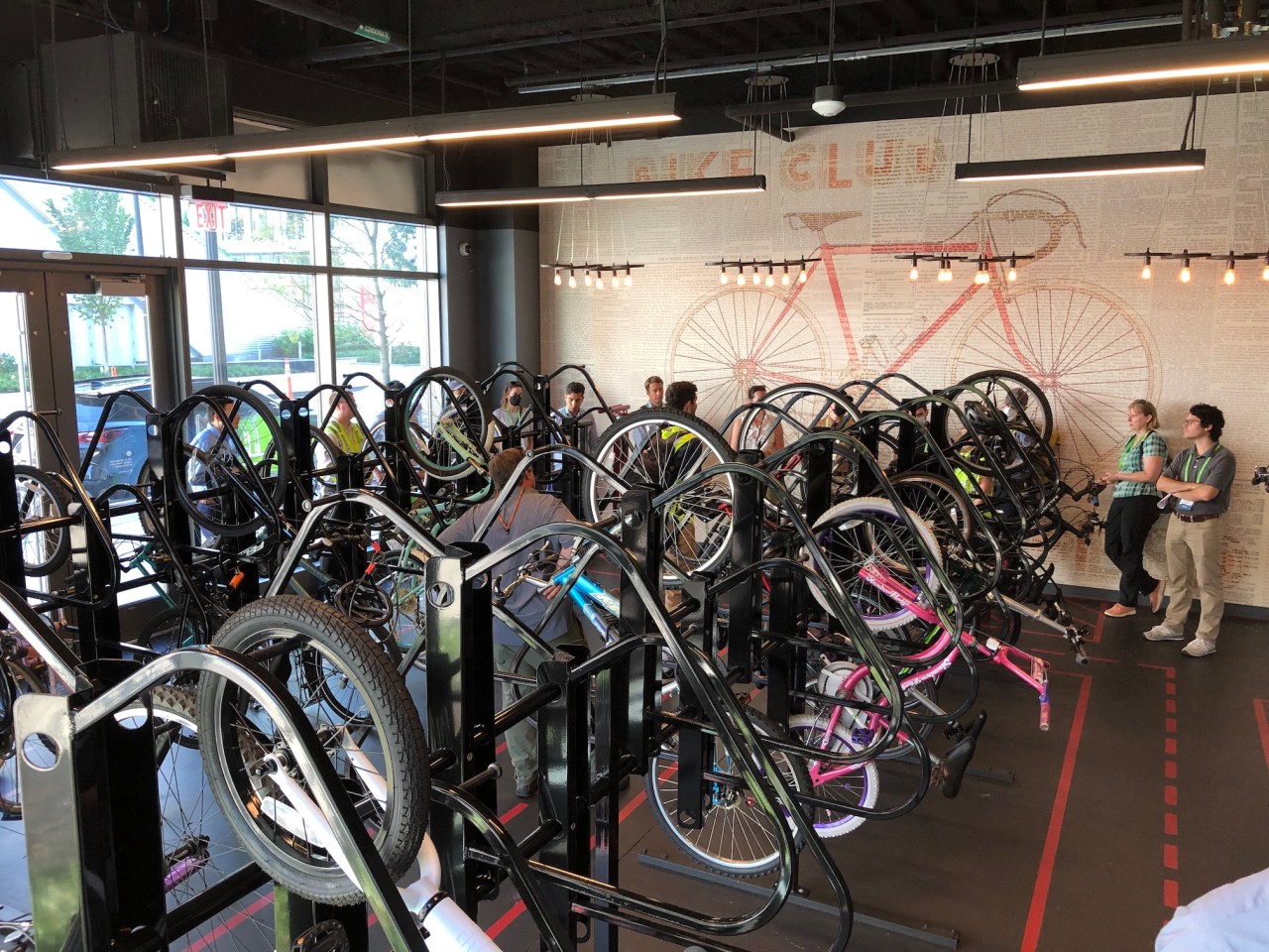 A room full of bikes hanging from vertical racks. The back wall features a mural of a bicycle and the wall to the left is full of windows looking out onto the sidewalk, including an exterior door.