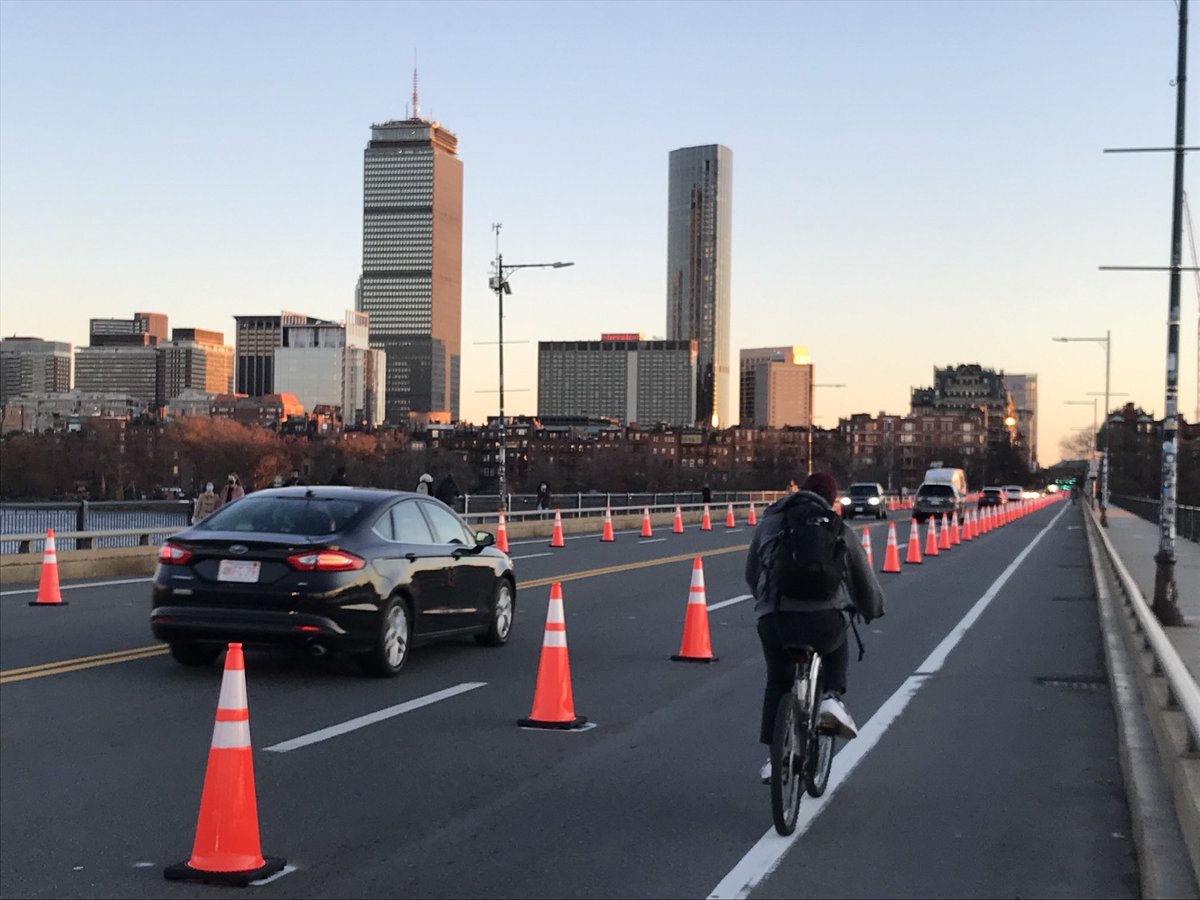 A row of cones delineates a bike lane on the Mass. Ave. bridge. A bicycle rider coasts by on one side of the cones while a car drives by to the left. On the horizon are the skyscrapers of the Back Bay neighborhood reflecting the late-afternoon sunlight.
