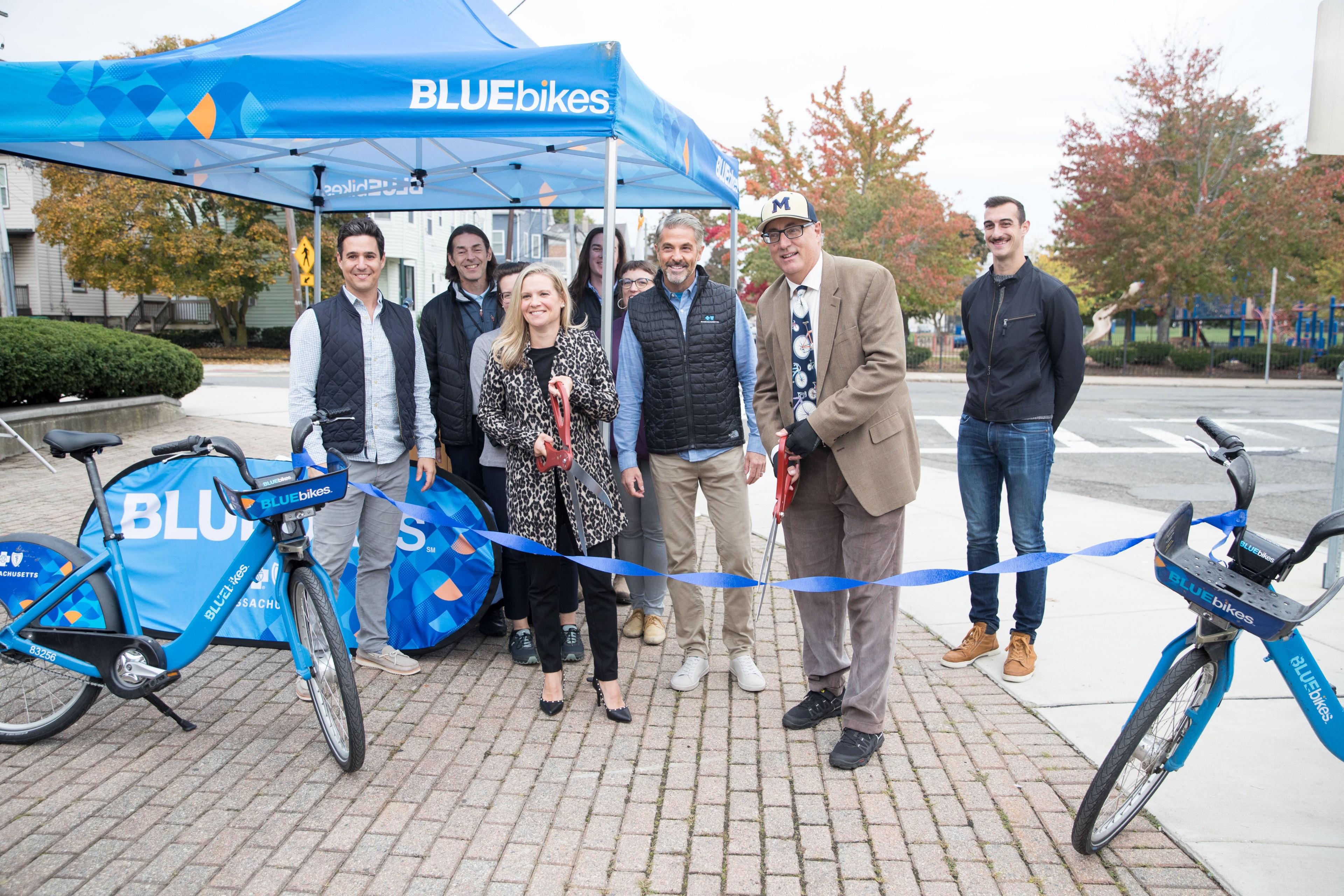 A small crowd gathers under a Bluebikes-branded pop-up tent next to some Bluebikes resting on their kickstands. In the foreground, a man in a suit, ballcap, and bicycle-print tie and woman in a leapord-print jacket hold comically oversized shears as they cut a ribbon while the others look on.