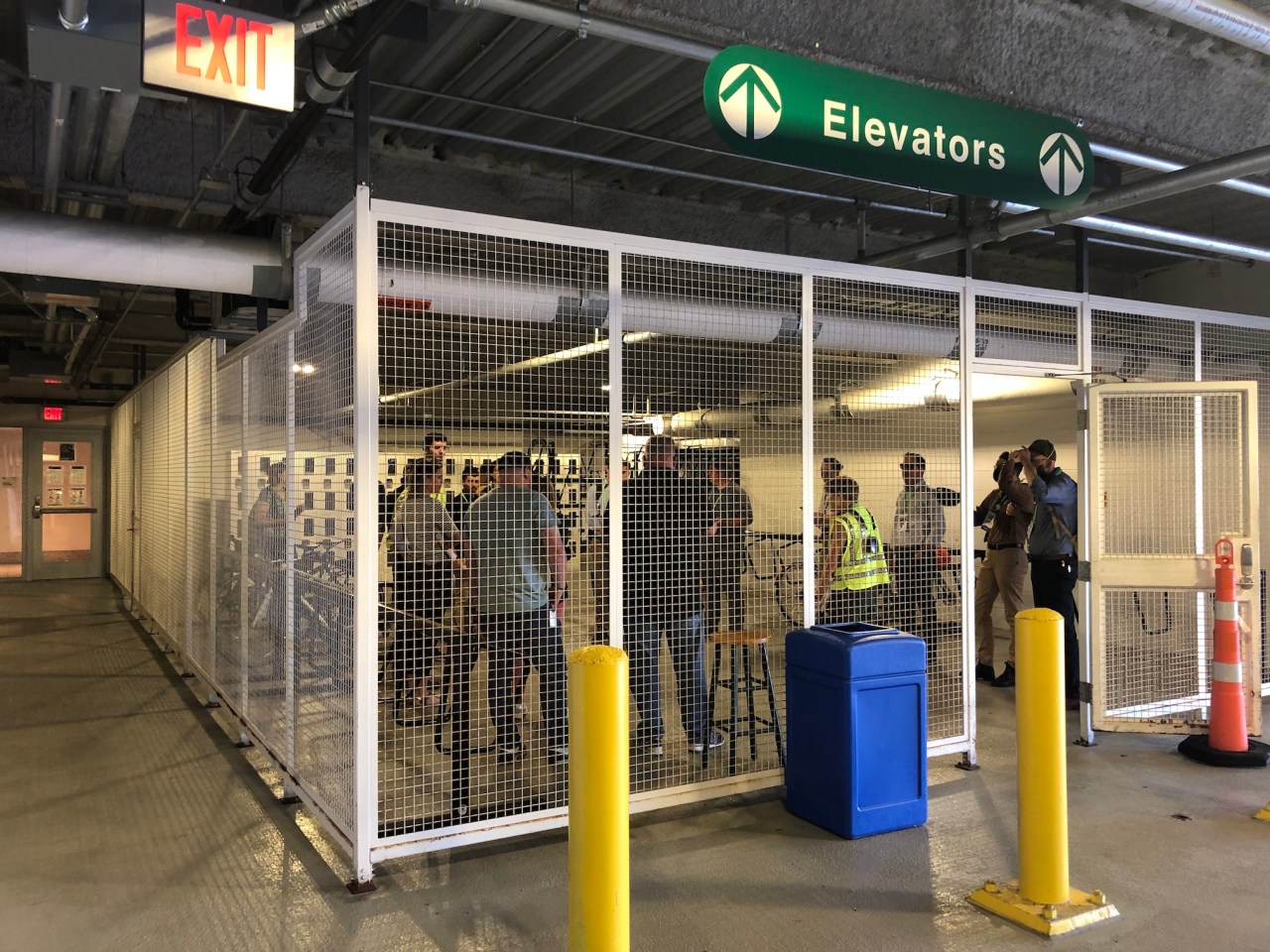 A small crowd chats inside a bike parking area surrounded by a white chain-link fence in a parking garage. "Exit" signs on the ceiling above point to a door in the distance next to the bike parking area, which leads to the office building's lobby. The back wall of the bike storage room is covered with lockers.
