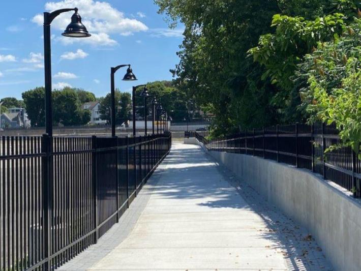 A new concrete shared-use path runs alongside fencing with lights towards a busy roadway in the distance. To the right of the path are some trees, and to the left, beyond the fence, is an asphalt parking lot.