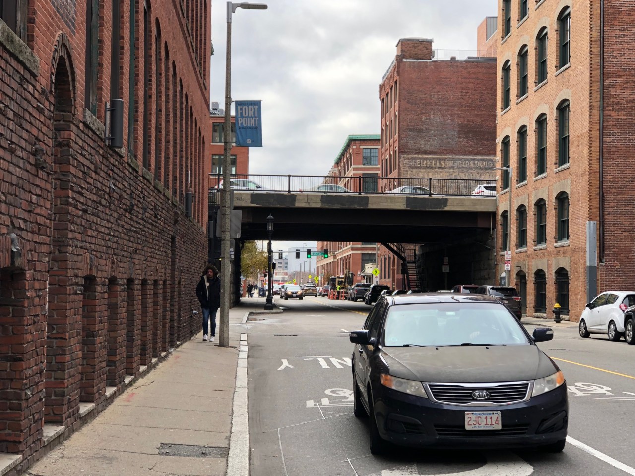 A city street with extremely narrow sidewalks obstructed by lightposts. Historic brick warehouse buildings line both sides of the street. In the distance is an viaduct carrying another street overhead.