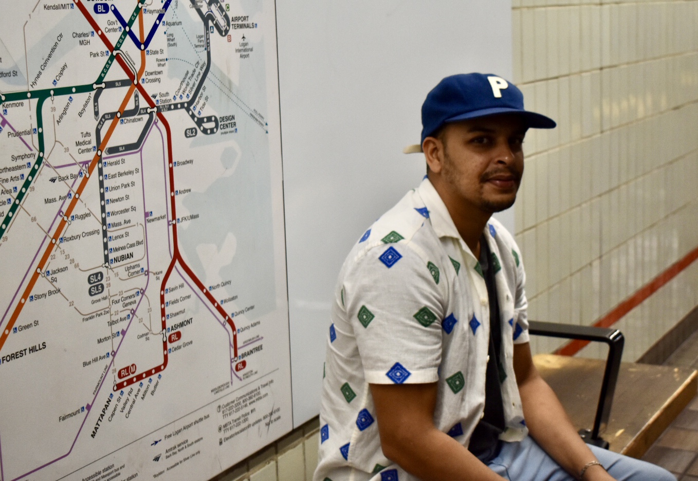 A man in a blue ballcap and white collared shirt sits on a bench in front of an MBTA rapid transit map inside a subway station.