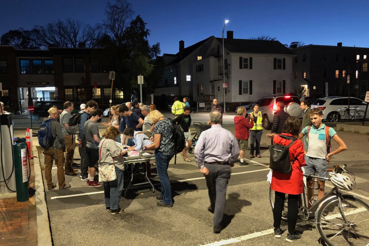 A crowd of people gathered in a parking lot to discuss plans for Hampshire Street. Most of the crowd is gathered around a table on the left side of the photo, where a large map of the street is on display. Other people talk with each other in small groups in other parts of the parking lot. Several attendees are carrying bikes or strollers.