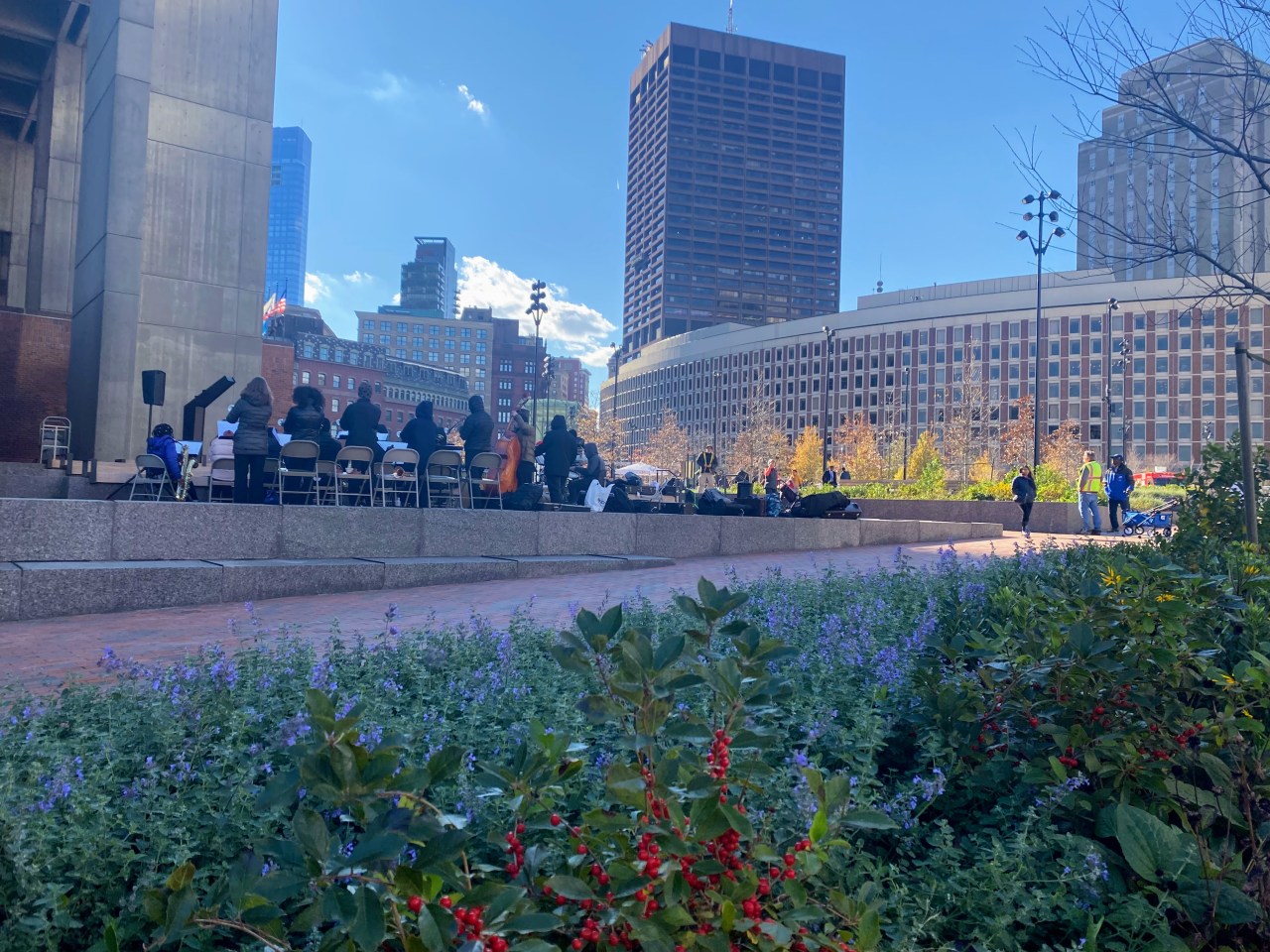 a student band plays behind blue and red flowers sitting among a bed of greenery on a portion of the plaza under a blue sky.