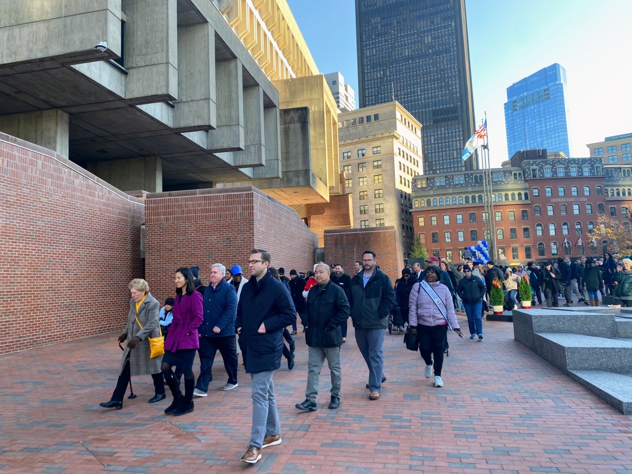 a group led by Boston Mayor Michelle Wu walks along the side of City Hall.