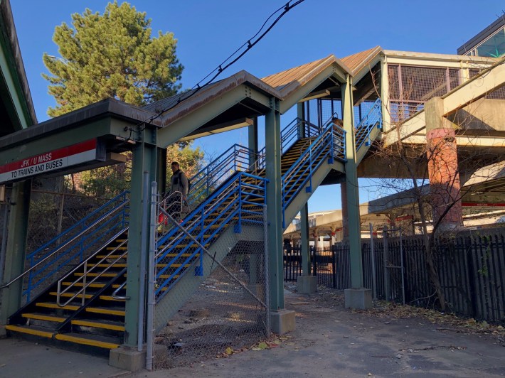 A covered stairway with three flights climbs up to a bridge over railroad tracks. A red sign above the ground-level entrance to the staircase reads "JFK/UMass - to trains and buses"