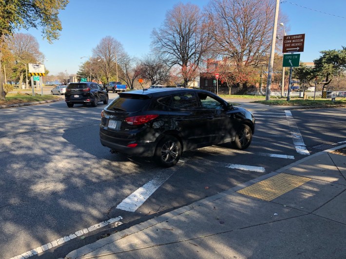 A black SUV drives through a painted crosswalk at the exit to a traffic circle. There are no traffic lights to stop traffic, only a yield sign facing the opposing lanes of traffic, where drivers enter the circle. A directional sign points to "Expo Center" to the left and "JFK Library/Comm Museum/State Archives" to the right.