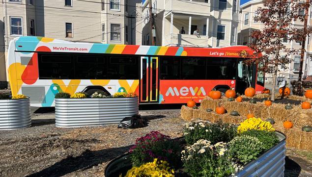 Riders check out a newly-rebranded Merrimack Valley Transit bus, with colorful red, blue, and orange stripes wrapped around this bus, next to a fall-themed park with haybales, mums, and pumpkins in Lawrence, Massachusetts. In the background are several triple-decker apartment buildings. Courtesy of Merrimack Valley Transit.
