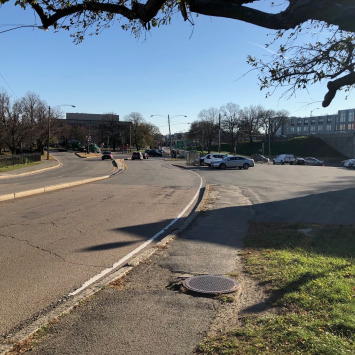 A broken, potholed asphalt sidewalk runs between a parking lot and a wide, mostly empty 4-lane roadway, both areas with much smoother pavement.