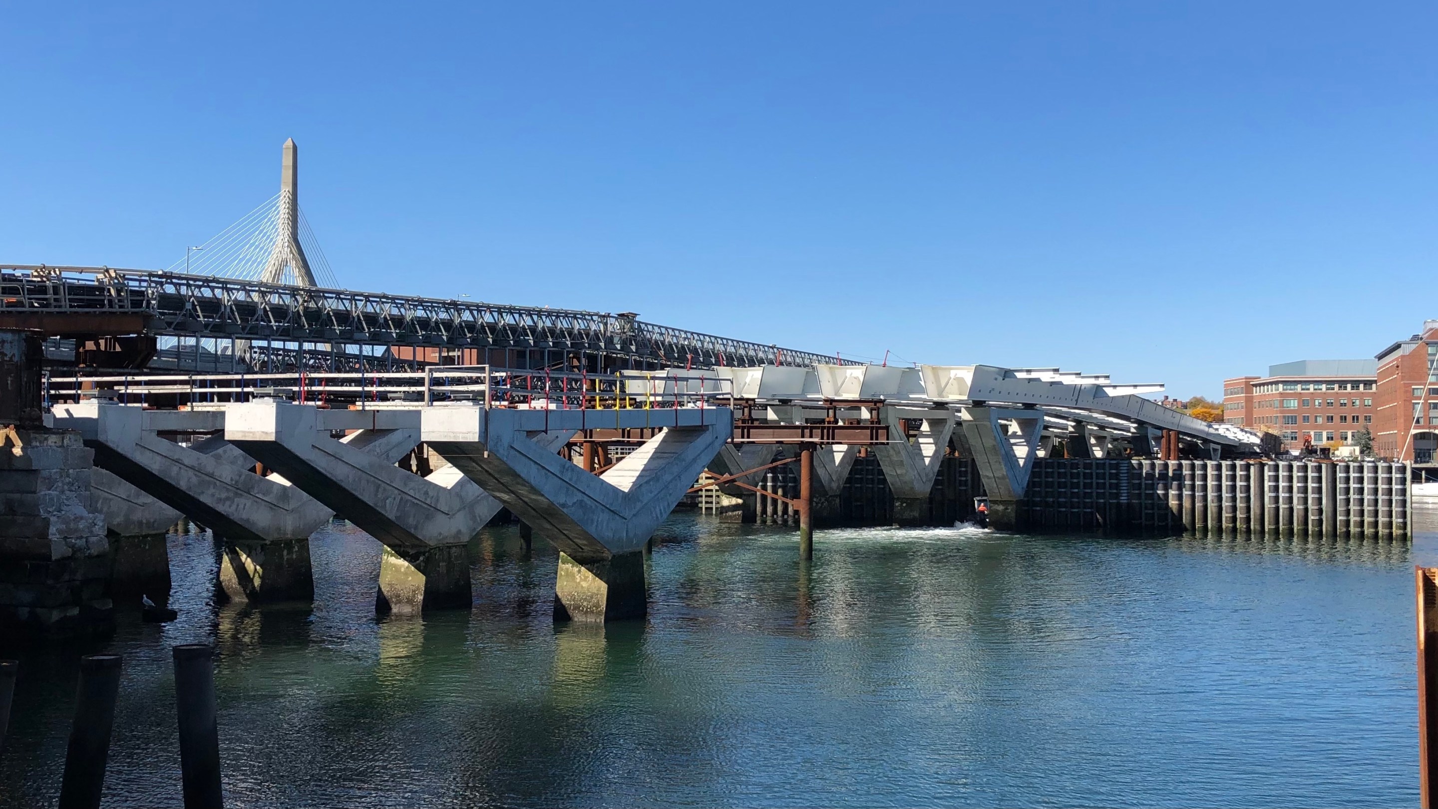 A bridge under construction. In the foreground are four piers, and behind them, large steel beams sit on top of another set of piers. The top of the cable-stayed Zakim Bridge is visible in the background.