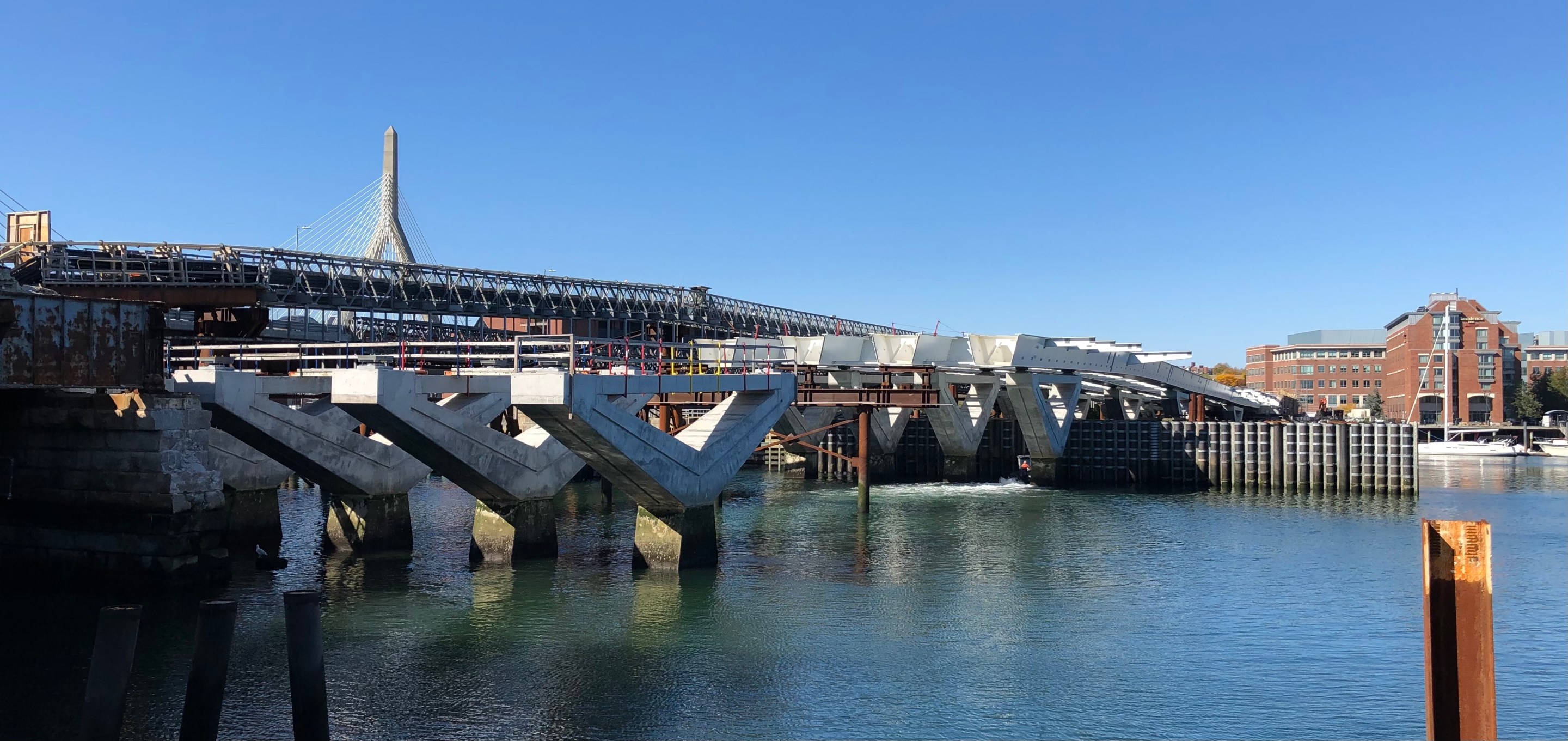 A bridge under construction. In the foreground are four piers, and behind them, large steel beams sit on top of another set of piers. The top of the cable-stayed Zakim Bridge is visible in the background.