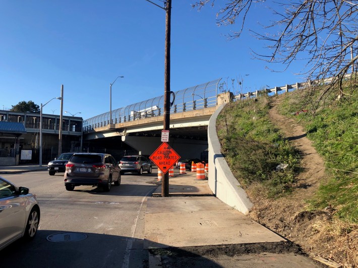 A highway overpass over a busy roadway. To the right, next to a retaining wall, a wide dirt path that's been trampled through the grass rises up from the lower roadway's sidewalk to the level of the overpass.