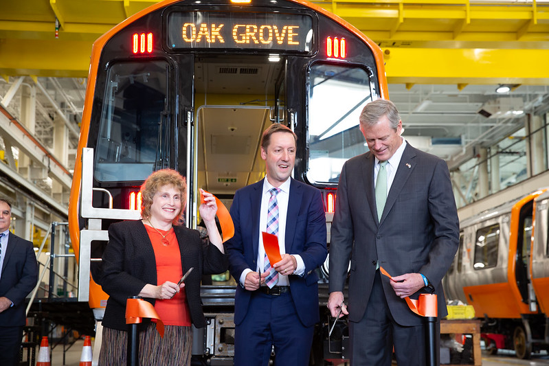 MassDOT Secretary Stephanie Pollack, wearing a blazer over a red top, stands next to Steve Poftak (center) wearing a suit and pink-and-blue plaid tie, and Mass. Gov. Charlie Baker, wearing a buttoned suit and green tie. All three are holding scissors and segments of a cut orange ribbon. They stand in front of a new Orange Line train car bearing the words "OAK GROVE" on the destination sign above the door.