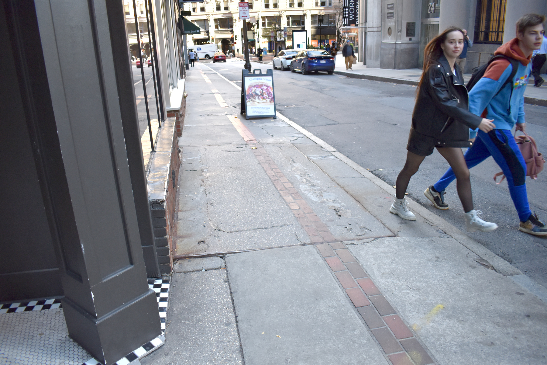 A man and an woman step off the curb on a sidewalk in downtown Boston. A row of red bricks running down the middle indicate the path of the Freedom Trail. In the center of the sidewalk is a large rectangular hatch that leads to the basement underlying the sidewalk.