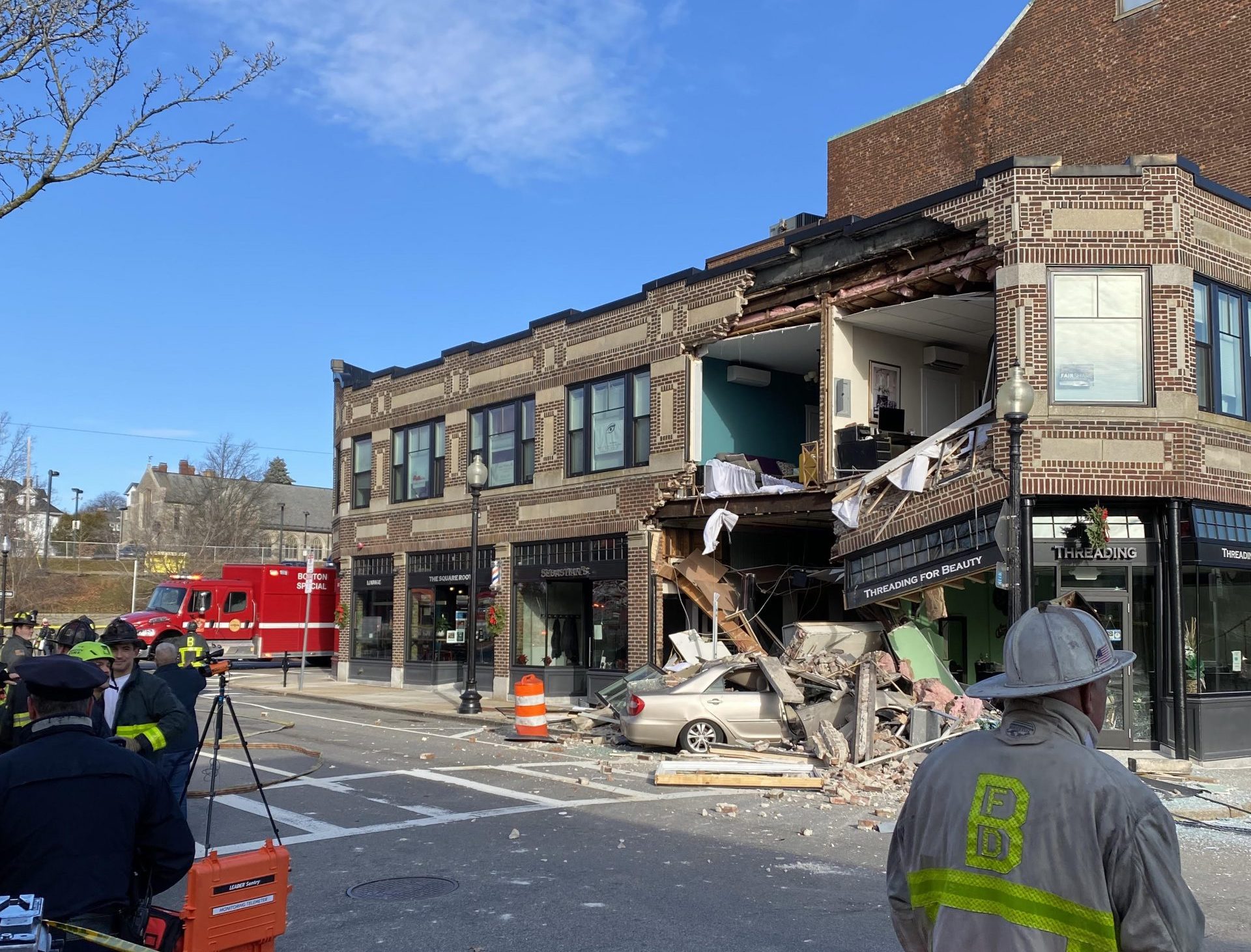 The aftermath of a car crash. A gold-colored Toyota sedan is buried in rubble with its front end poking into a barbershop on the ground floor of a two-story brick building with several small business storefronts. Above it, a section of the building's wall is completely collapsed, leaving second-floor offices exposed to the open air. Several firefighters mill about in the foreground across the street.