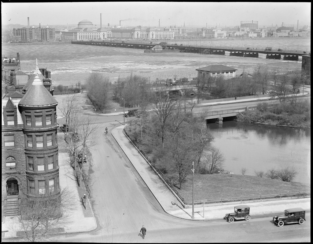A historic black-and-white photograph of Charlesgate Park before the Bowker Overpass and Storrow Drive existed. The photo is taken from an elevated vantage point looking down at the Muddy River and the bridges that carry Commonwealth Avenue, Beacon Street, and Back Street over it. In the foreground two Model T-type cars drive along a mostly empty Commonwealth Avenue while a man crosses the street on foot in front of them. In the background is the Charles River and the Harvard Bridge, and the Great Dome of MIT is visible on the other side of the river in a mostly industrial riverfront district in Cambridge.
