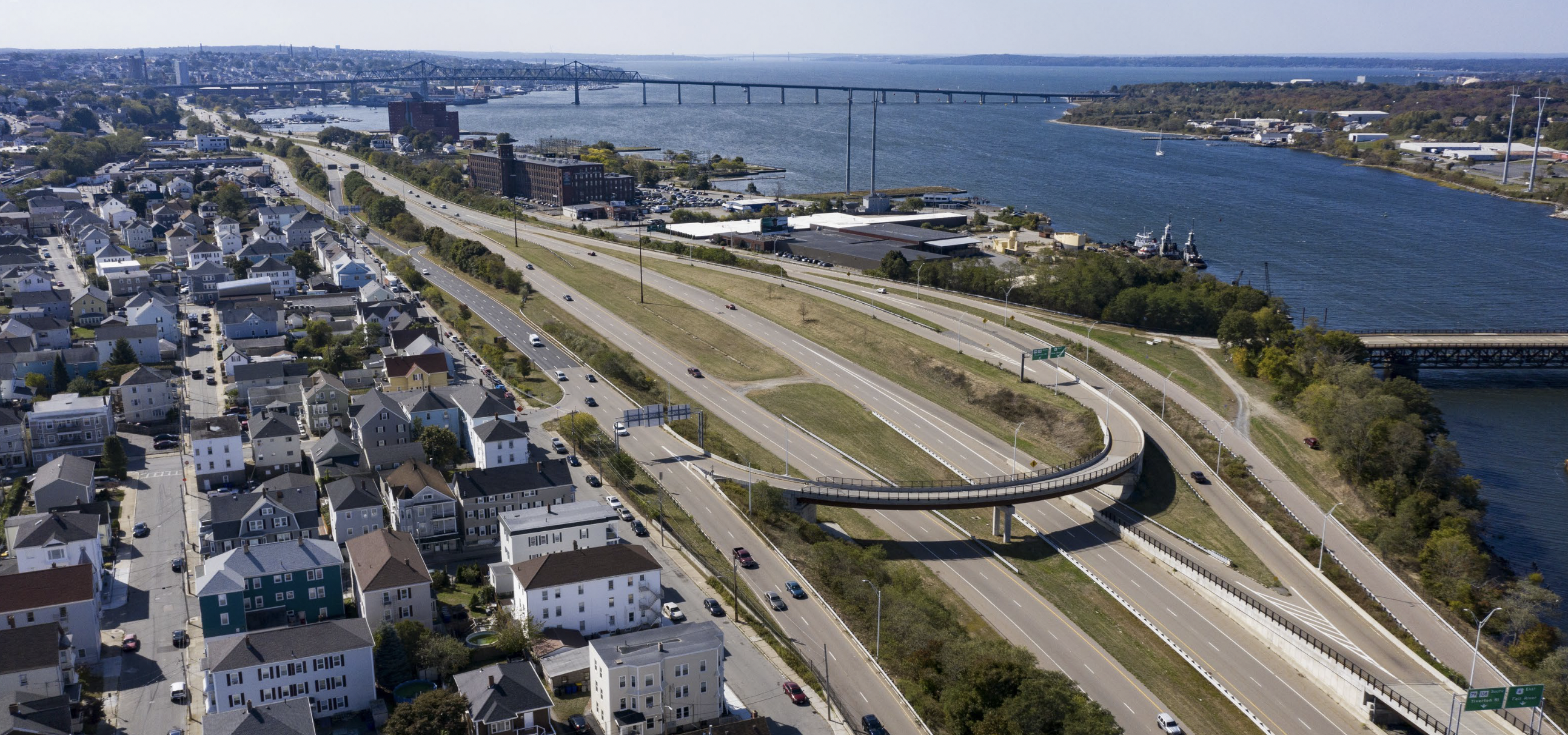 An aerial view of Fall River's Route 79 expressway, looking south towards downtown Fall River (top left) and Mt. Hope Bay (top right). The view is dominated by the massive Route 79, which occupies 8 to 14 lanes alongside wide expanses of empty grass along Fall River's waterfront. Along the left edge of the photo is visible the densely-populated residential neighborhoods of Fall River's North End.