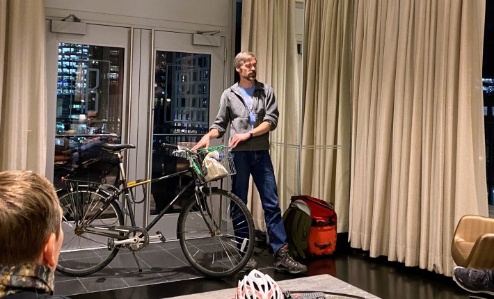 Galen Mook stands next to his black steel winter bike infront of a set of doors besides tall tan curtains