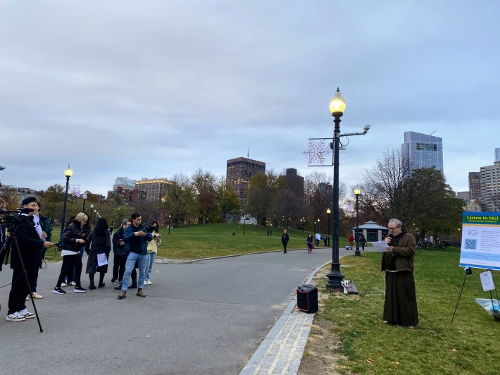 An older man wearing a Franciscan monk's habit speaks next to a poster-sized display board with the title "Listen to Me! Stories of the unnoticed" on the edge of a paved pathway in Boston Common. To his left are a small crowd of people with cameras and other recording equipment. Downtown high-rises are visible on the horizon under grey skies.