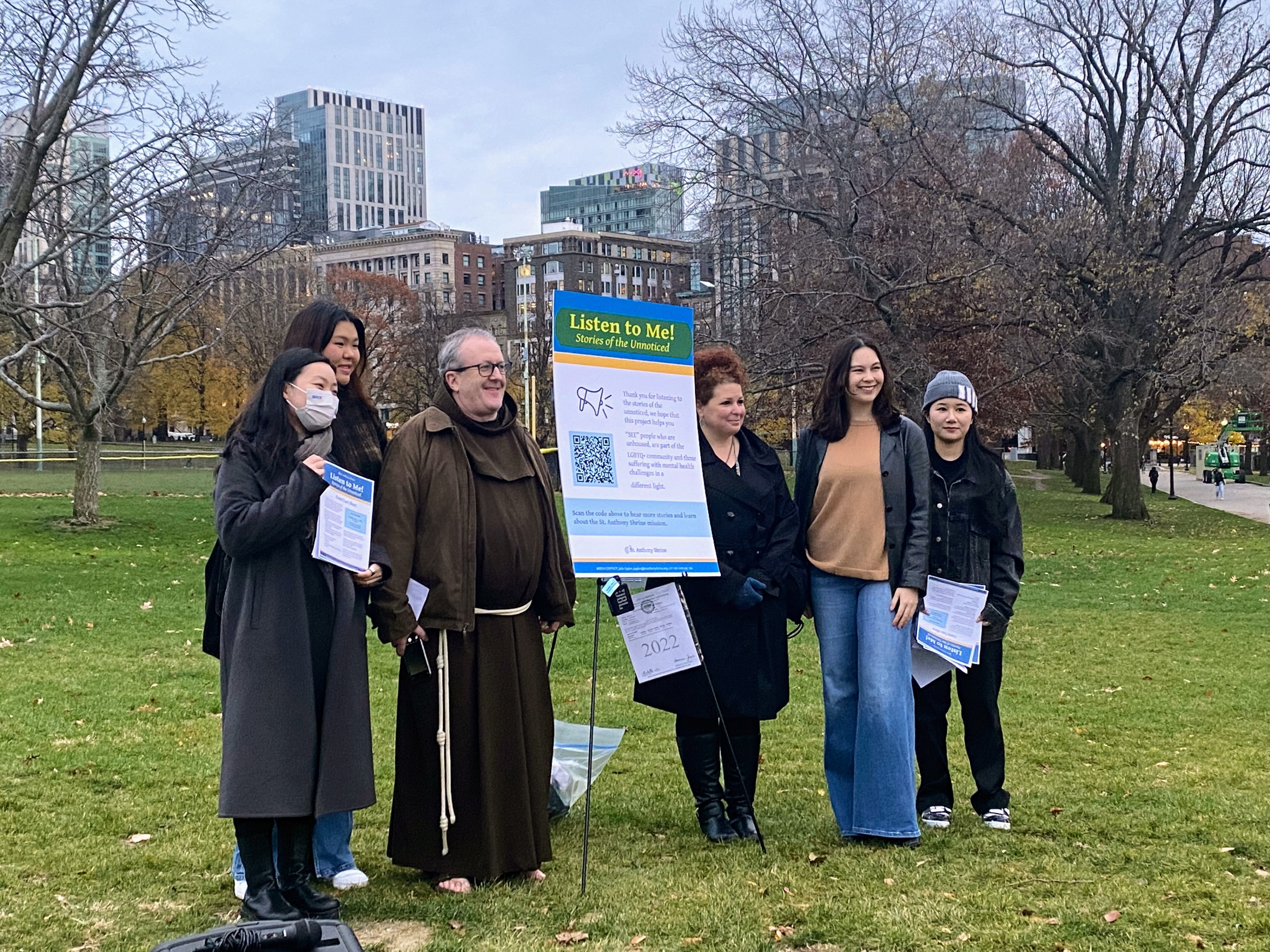 Five college students in winter clothes stand around an older man wearing a Franciscan monk's habit, and a poster-sized display board with the title "Listen to Me! Stories of the unnoticed" on a grassy lawn in Boston Common. Downtown high-rises are visible on the horizon under grey skies.