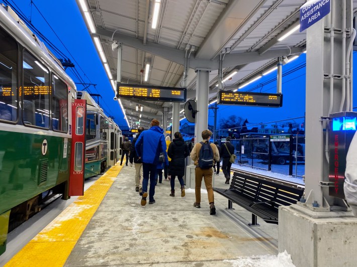 Passengers walk along a snow-covered platform next to a stopped Green Line train. Signs indicate that this is Ball Square station.