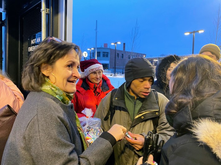 Somerville Mayor Katjana Ballantyne, wearing a gray winter coat, passes out souvenir buttons to a crowd of MBTA passengers, also wearing winter overcoats.