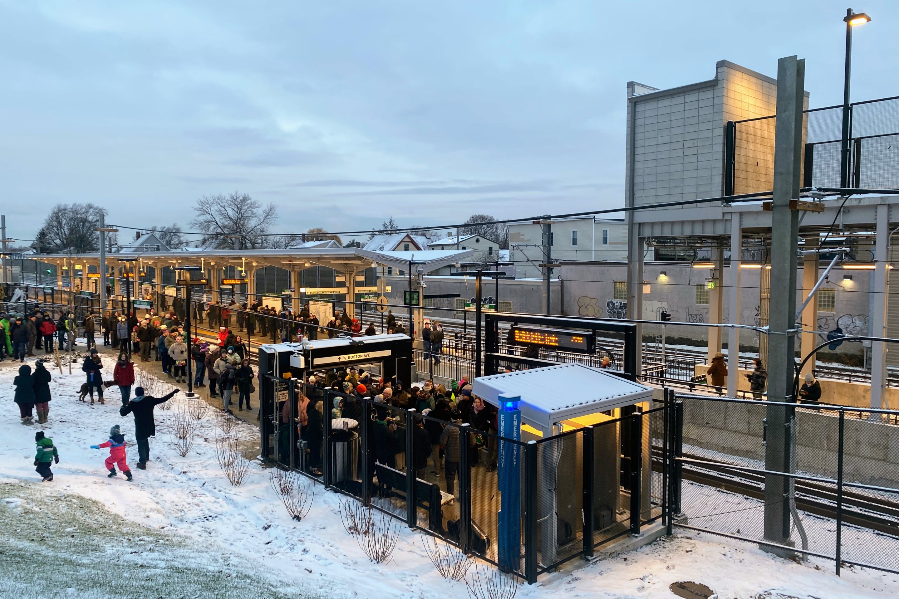 A large crowd of people celebrate the Green Line extension's opening day outside a snowy Ball Square station while some kids play on a snowy hillside next to the station entrance.