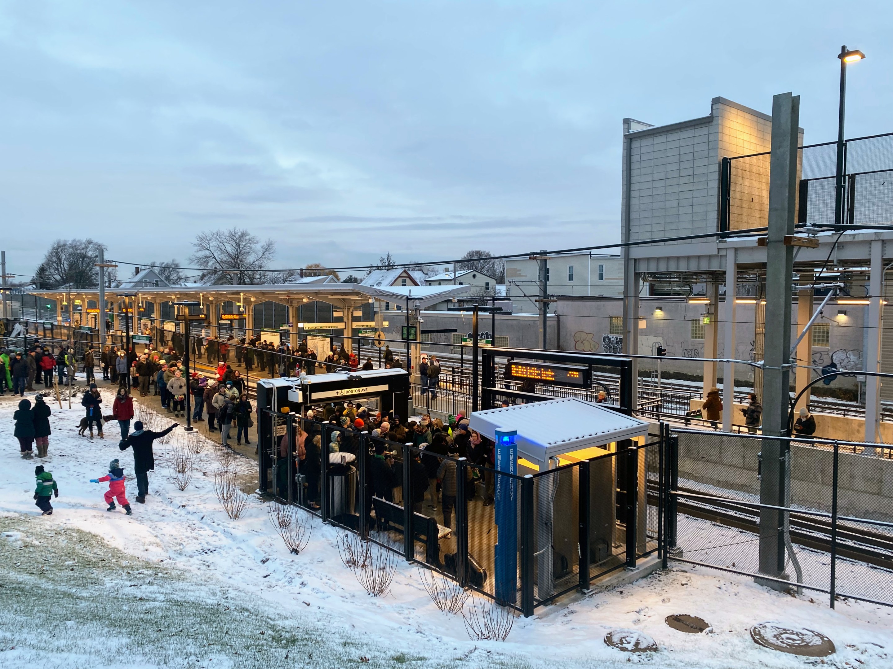 A large crowd of people celebrate the Green Line extension's opening day outside a snowy Ball Square station while some kids play on a snowy hillside next to the station entrance.
