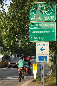 A green bike route wayfinding sign in Provincetown, next to a tree-lined street where two people on bikes are riding. A rectangular sign on top of the signpost has a white image of a bike above the words "BIKE ROUTE" next to a an outline of Cape Cod, on a green background. Below, another green-background sign split into three sections shows how to get to various destinations: a leftward arrow points to "Old Colony Path," a rightward arrow points to "Commercial Street," and a straight arrow points to "Truro".