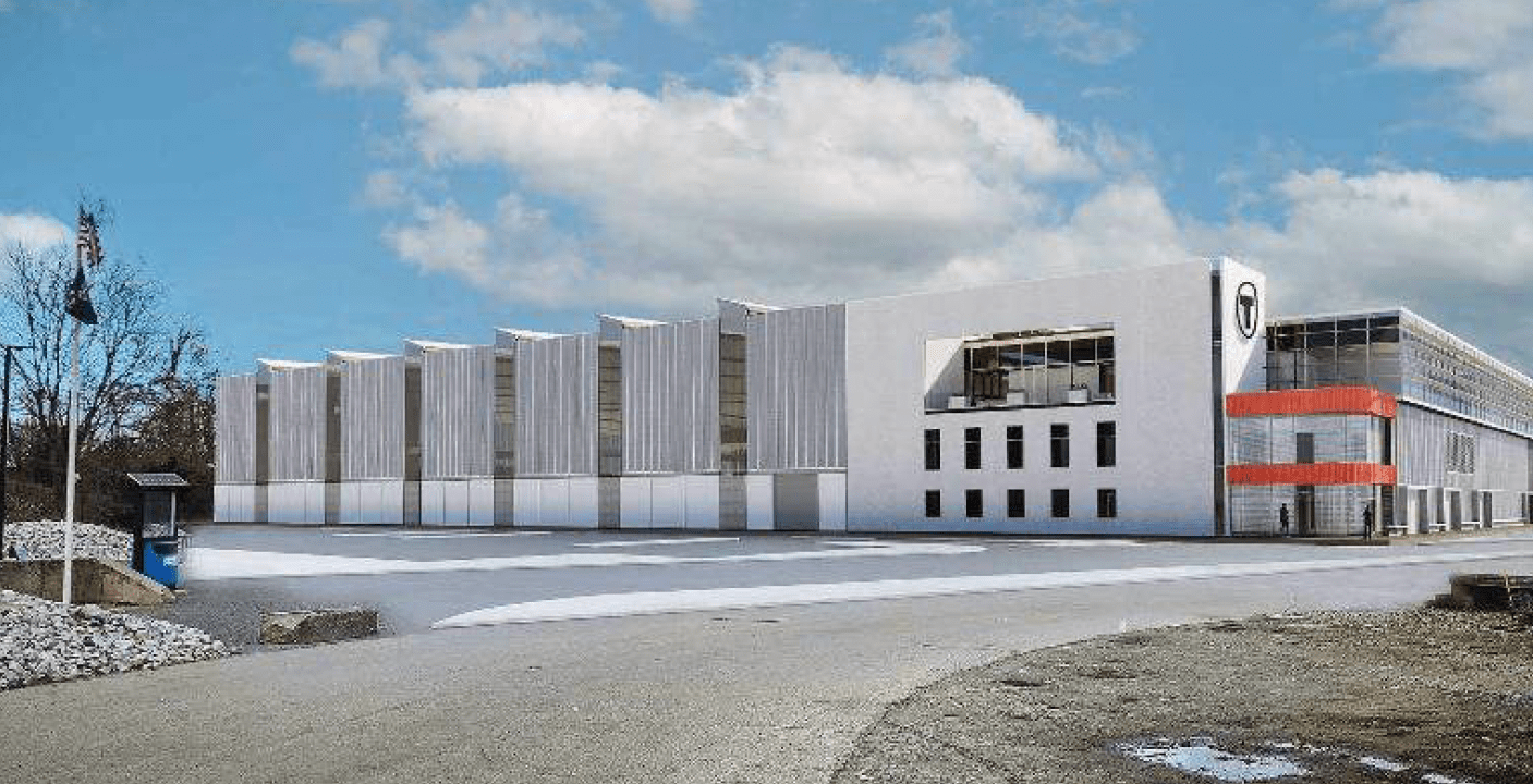 A rendering of the MBTA's proposed new South Side Commuter Rail Maintenance Facility, a large grey-and-white warehouse building. The MBTA's T logo is located on the corner of the building above an entrance doorway.