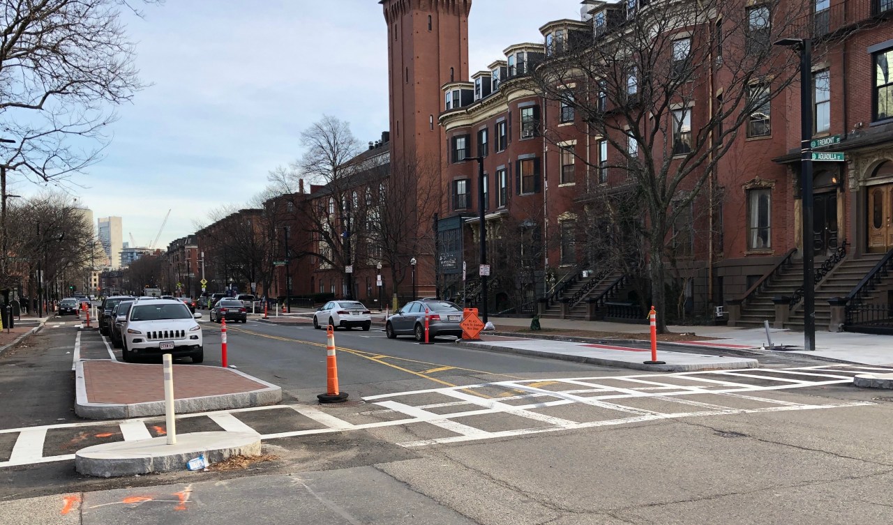 A new crosswalk on Tremont Street in the South End. In the foreground are four newly-installed median islands paved in bricks and granite. The islands separate the street's bike lanes, which run along the edge of the street, separated from the center car lanes by the islands at the intersection and rows of parked cars further in the distance. Historic brick rowhouses line the opposite side of the street. In the distance on the left some skyscrapers and construction cranes in downtown Boston are visible. 