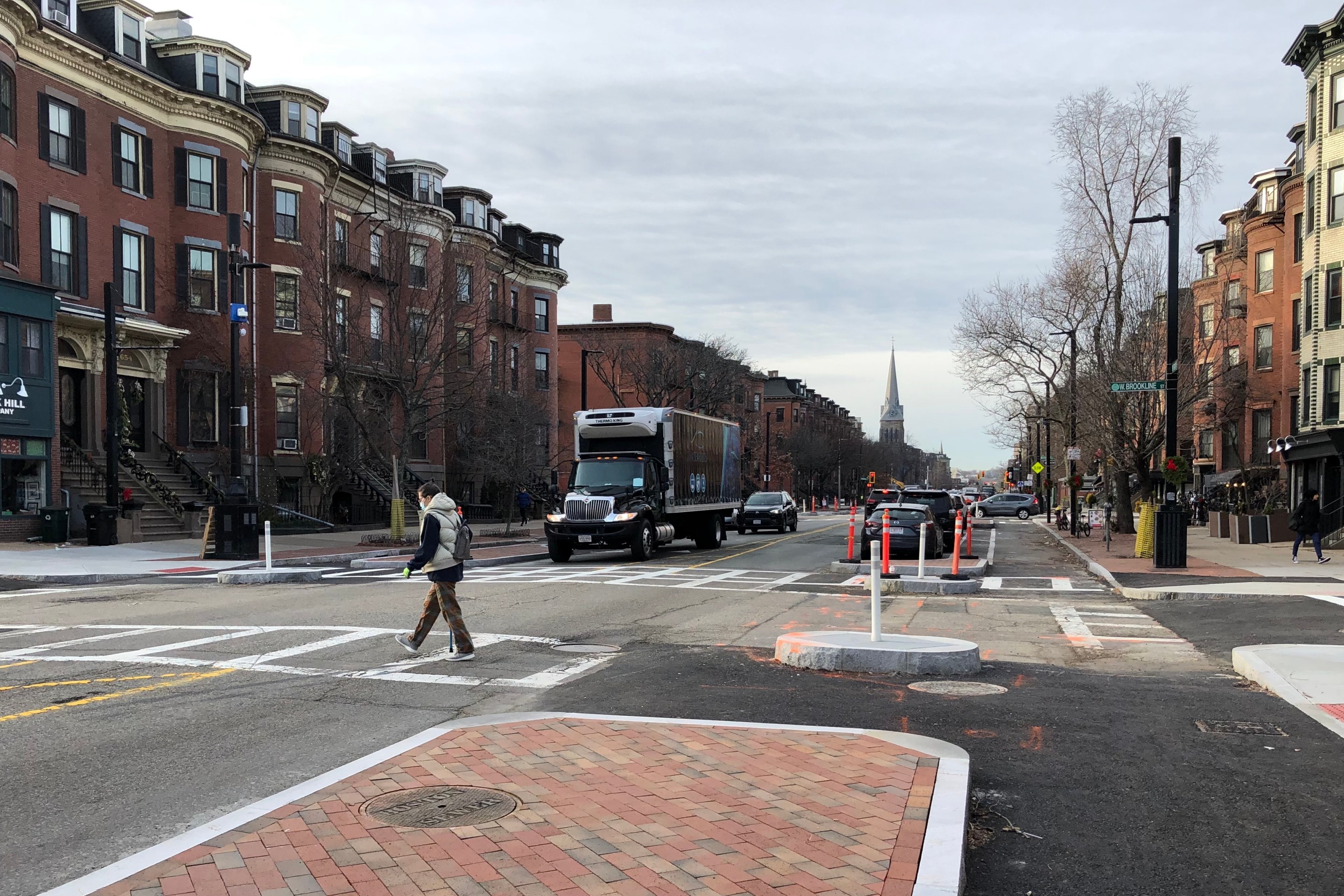 A pedestrian crosses Tremont Street in a new crosswalk. In the foreground is a newly-installed median island, about the size of a parked car, that is paved in bricks and ringed by a granite curb. To its left are two lanes for two-way car traffic; to its right is an asphalt-paved bike lane, and a second curb at the right edge of the photo delineates the street's sidewalk. A smaller granite curb island is located at the far side of the crosswalk, and two other pairs of islands in a similar configuration protect the crosswalk on the opposite side of the intersection. Historic brick rowhomes line both sides of the street. In the distance is a church steeple.