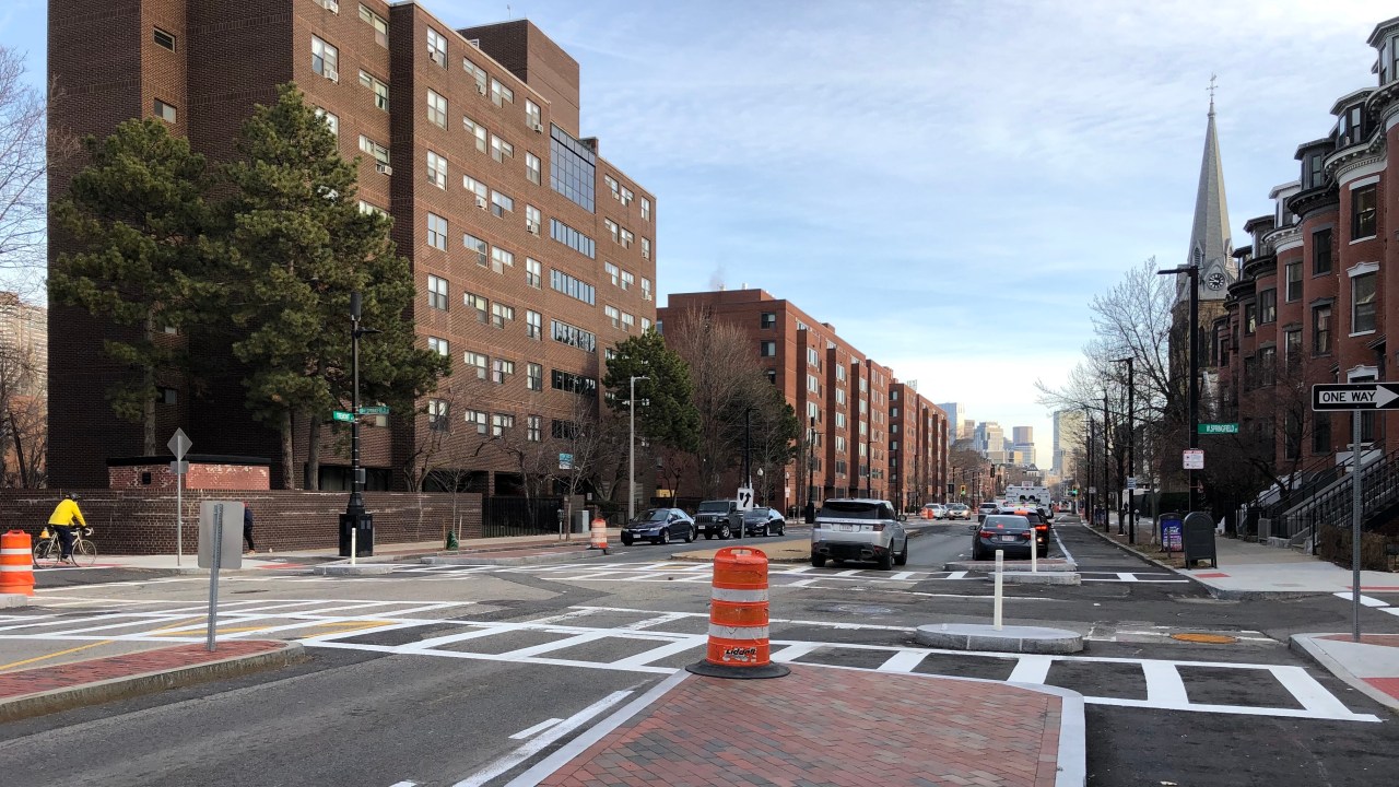 A new crosswalk on Tremont Street in the South End. In the foreground is a newly-installed median island, about the size of a parked car, that is paved in bricks and ringed by a granite curb, with an orange construction barrel on top. To its left is a single motor vehicle lane and another, smaller median island; on the left edge of the photo, a person wearing a bright yellow jacket rides a bicycle across the street. On the far side of the intersection are three more median islands to delineate traffic: an older concrete median in the middle of the street divides eastbound and westbound motor vehicle traffic, and smaller, newly-installed medians paved in brick and granite on either side of the street separate the car lanes from bike lanes, which run along the street's sidewalks. Beyond the median islands, the bike lanes are separated from car lanes by rows of parked cars. Two modern, brick-clad 20th-century apartment blocks line the opposite side of the street. In the distance on the right is a stone church steeple and clocktower.