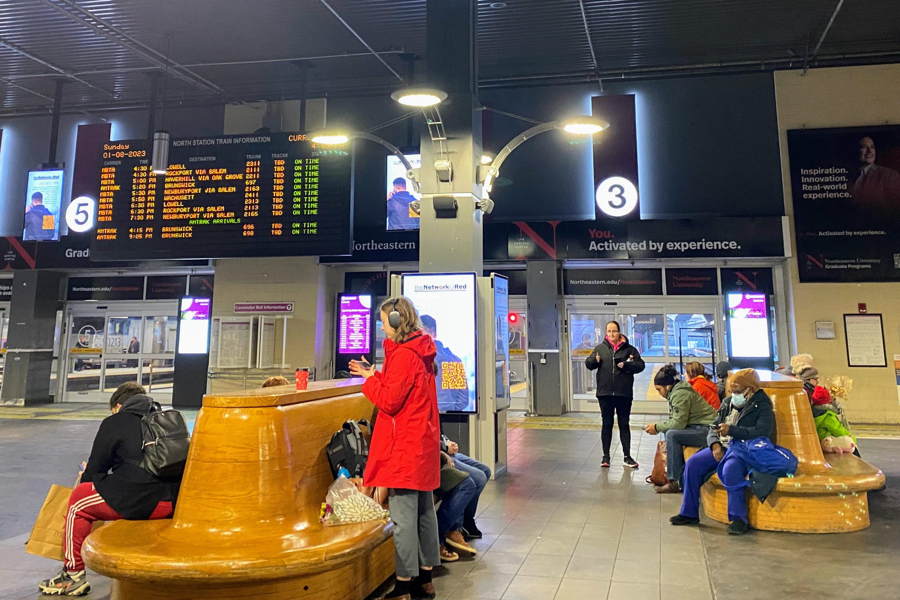 folks stand or sit on large wooden benches while waiting for their train inside North Station. The timetable hangs high in the background.