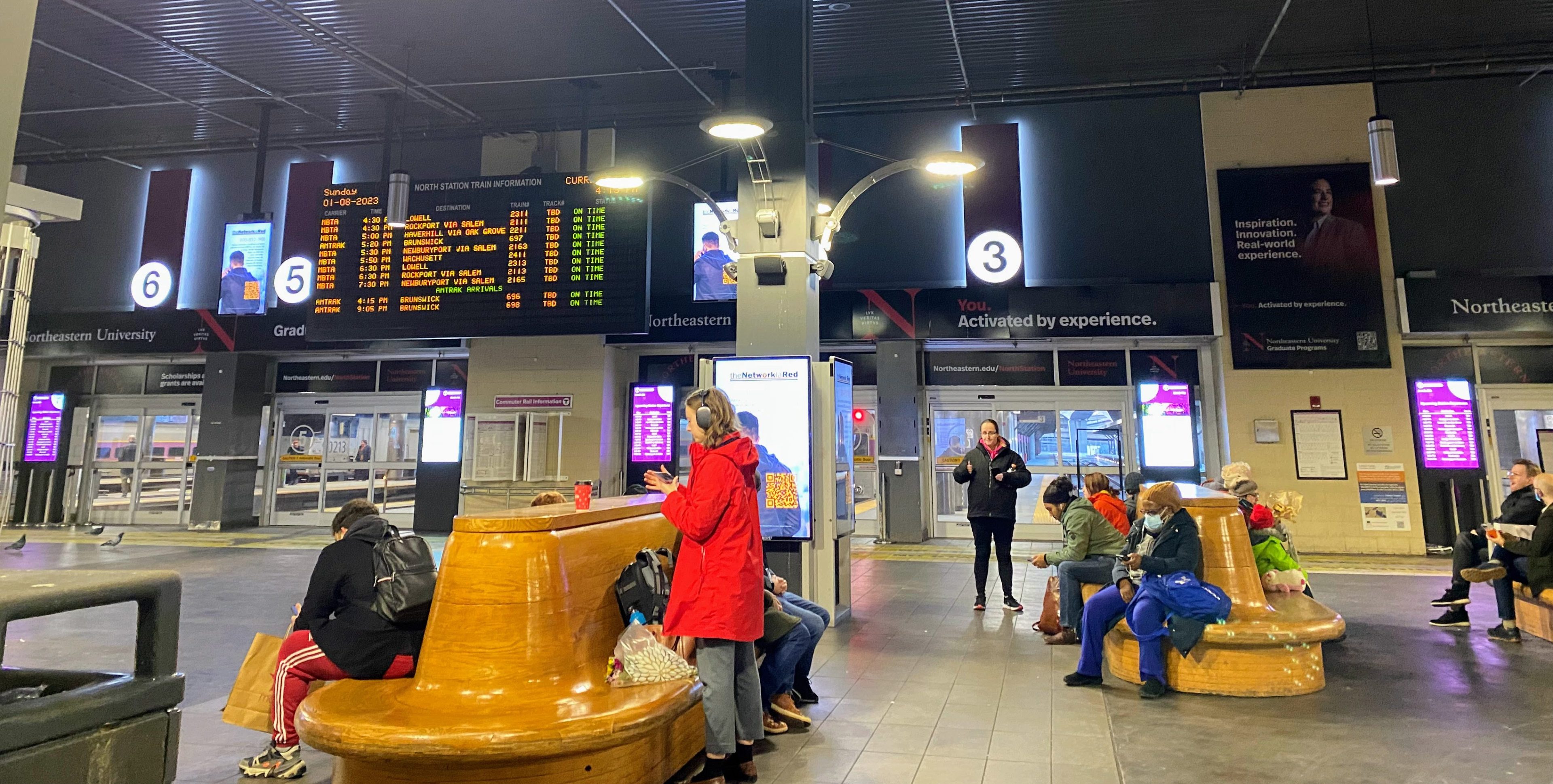 folks stand or sit on large wooden benches while waiting for their train inside North Station. The timetable hangs high in the background.