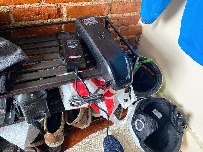 an cylindrical shaped e-bike battery charges on top of a shoe rack up against a brick wall.