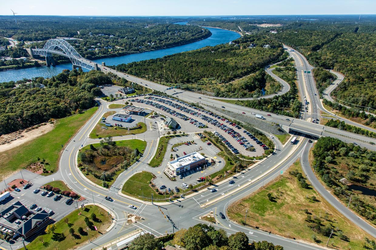 An aerial view of a major highway interchange. In the foreground is the intersection of two multi-lane suburban roads. Behind it is a large parking lot, with a gas station and a handful of other highway-oriented businesses surrounded by asphalt. In the middle distance, a four-lane expressway runs from the right to upper left. In the upper left, the highway crosses a canal over an arched bridge. Another highway runs parallel to the canal into the distance in the photograph's upper right corner.